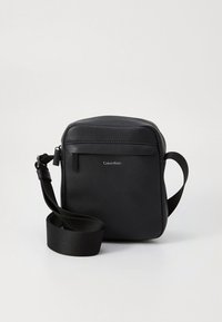 Black Calvin Klein crossbody bag with adjustable strap, textured finish, front zipper pocket, and silver logo on plain background.
