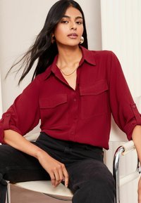 Chemisier rouge avec un devant boutonné, deux poches poitrine et des manches retroussables. Tissu lisse et léger avec une coupe décontractée. Jean noir.