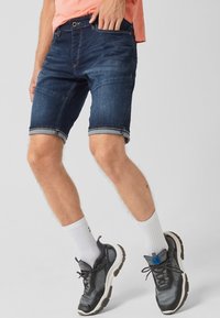 Shorts de mezclilla en azul oscuro con dobladillos enrollados, con bolsillos delanteros y un diseño ajustado, combinados con zapatillas oscuras y calcetas blancas.