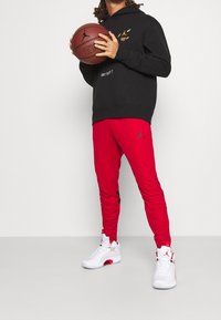 Sweat à capuche noir avec texte en or et blanc, pantalon de jogging rouge avec accents noirs, et baskets de basketball blanches avec détails rouges.