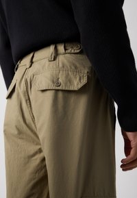 Beige cargo nohavice so bočnými vreckami, textúrovaná látka a zapínacie vrecká na zadnej strane. Nosené s čiernym rebrovaným sveterom, ktorý odhaľuje pás.