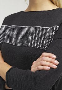 Pull en tricot noir avec un empiècement texturé en motifs blancs et noirs. Manches longues, col rond et texture douce.