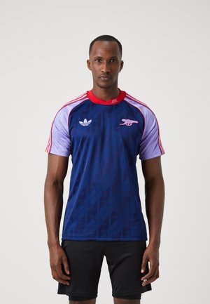 Maillot à manches courtes bleu et violet avec des accents rouges et un col rond. Présente les logos d'Adidas et d'Arsenal. Motif subtil sur l'ensemble.