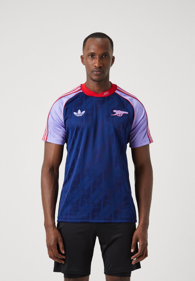Maglietta a maniche corte blu e viola con dettagli rossi e colletto rotondo. Presenta i loghi di Adidas e Arsenal. Motivo sottile su tutta la superficie.