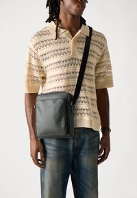 Borsa a tracolla in pelle grigia con tasca frontale, tracolla nera regolabile e texture liscia, indossata a crossbody sopra una camicia beige a fantasia.