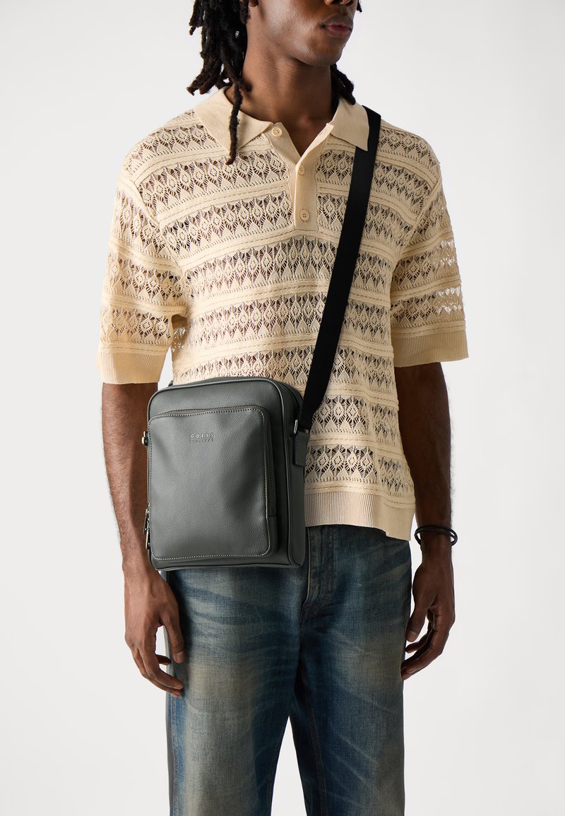 Borsa a tracolla in pelle grigia con tasca frontale, tracolla nera regolabile e texture liscia, indossata a crossbody sopra una camicia beige a fantasia.