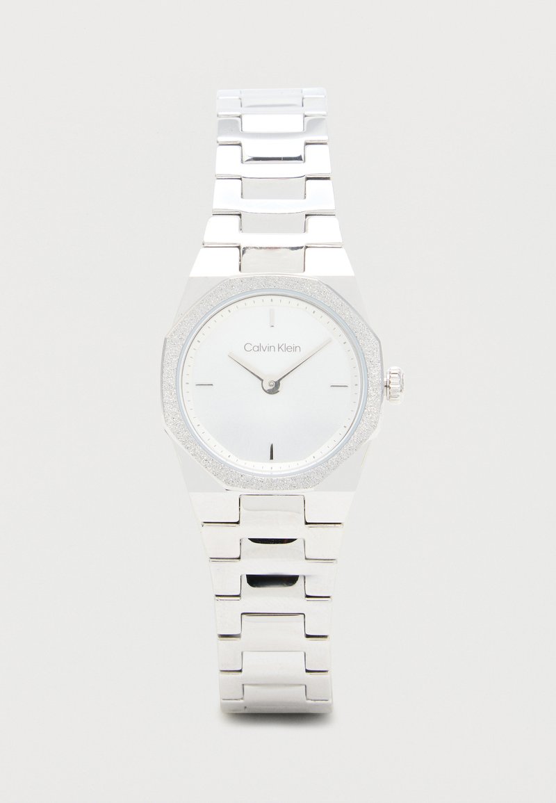 Montre-bracelet en argent avec un boîtier octagonal, un cadran blanc, des marqueurs d'heure minimalistes et une lunette scintillante ; présente une finition en métal poli.