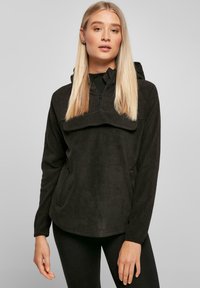 Maglione in pile nero con zip a metà davanti, cappuccio e tasca a marsupio. Presenta maniche lunghe e un materiale morbido e testurizzato.