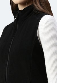 Schwarze Fleece-Weste mit hohem Kragen und Reißverschluss auf der Vorderseite. Strukturierter Stoff, figurbetont geschnitten, getragen über einem weißen, gerippten Langarmoberteil.