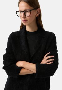 Černý, huňatý blejzr z texturované látky s klopovým límcem a manžetovými rukávy, vrstvený přes černý top s crewneck výstřihem.