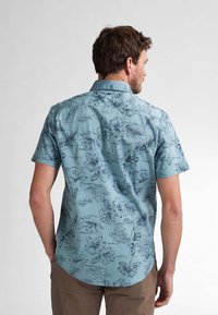 Camisa de manga corta con botones en tejido azul claro, que presenta un patrón de flores y paisajes en tonos de azul más oscuro. Dobladillo curvado.