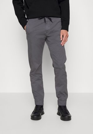 Mann trägt graue Kordelhose mit elastischen Bündchen und schwarze Schnürstiefel, steht mit entspannten Händen.