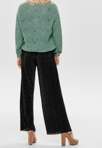 Mintgrüner, gestrickter Pullover mit Wellenmuster, kombiniert mit lässigen, schwarzen Hosen mit Polka-Dots und beigen Plateauschuhen.