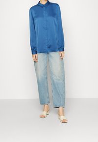 Blå satin långärmad skjorta med spetsig krage, knappframsidan, i kombination med ljusblå avslappnade jeans och beige öppna klackar.