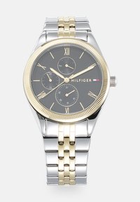 Montre-bracelet argentée et dorée avec un cadran noir, des chiffres romains, trois sous-cadrans et un bracelet en métal bicolore. Accent de lunette striée dorée.