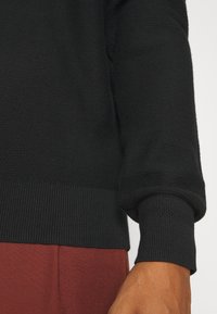 Gros plan sur le bras d’une personne portant un pull noir texturé avec des poignets côtelés et un pantalon marron.