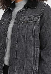 Černá džínová bunda s texturovaným límcem, zapínáním na knoflíky a kapsami na prsou. Má kontrastní prošití a klasický střih.