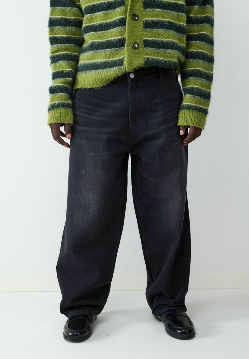 Spodnie dżinsowe w szerokim kroju, czarne, o gładkiej fakturze, z widocznym szyciem i dopasowanym paskiem w talii, zestawione z zielono-czarnym, prążkowanym swetrem dzianinowym.