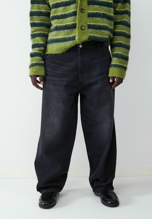 Wijd uitlopende zwarte denim jeans met een gladde textuur, voorzien van zichtbare stiksels en een aansluitende tailleband, gecombineerd met een groene en zwarte gestreepte gebreide trui.