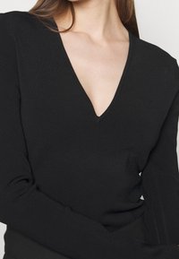 Svart långärmad topp med djup V-ringning, ribbad textur och figurnära silhuett. Slät tyg fortsätter ner till handleden.
