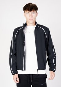 Giacca leggera nera con dettagli bianchi, chiusura con cerniera, colletto alto, polsini elastici e tasche laterali. Indossata sopra una t-shirt bianca.