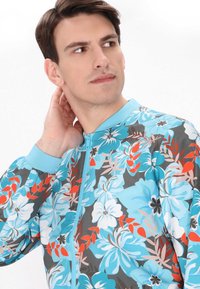 Giacca zip-up floreale in un vivace motivo blu-grigio, con grandi fiori bianchi e turchesi con accenti rossi e polsini blu.