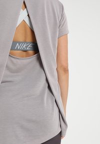 Top deportivo gris con un diseño holgado en la parte posterior, que revela un sujetador deportivo blanco de Nike con tirantes entrecruzados. Textura de tela suave.