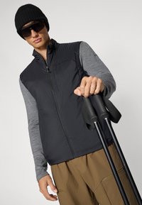 Zwarte lichte vest over een grijze lange mouwen shirt, bruine broek, zwarte beanie en zonnebril. Persoon houdt zwarte trekkingstokken vast.