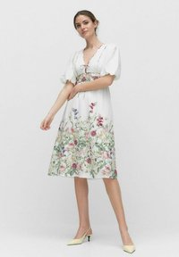 Weißes Blumenkleid mit Puffärmeln, einem V-Ausschnitt und einer taillierten Taille. Verziert mit bunten Blumenmustern am Rock. Getragen mit gelben High Heels.