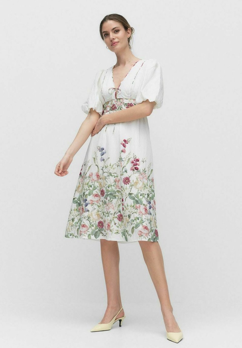 Weißes Blumenkleid mit Puffärmeln, einem V-Ausschnitt und einer taillierten Taille. Verziert mit bunten Blumenmustern am Rock. Getragen mit gelben High Heels.