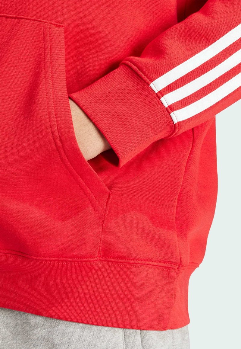 Sweatshirt rouge avec poche kangourou, poignets côtelés et trois bandes blanches sur la manche. Fabriqué en tissu doux avec une texture lisse.