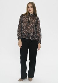 Blusa transparente de mangas largas con cuello alto y cremallera, en un patrón abstracto en marrón y negro, combinada con pantalones negros y zapatos adornados.