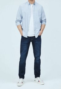 Randig skjorta i ljusblått, vit t-shirt under, mörkblå jeans och vita sneakers med färgade accenter.