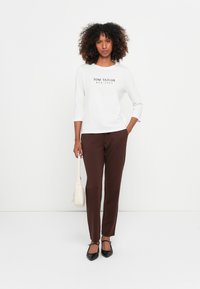 Witte shirt met "TOM TAILOR EST. 1962" in zwart gedrukt, gecombineerd met bruine broek. Accessoires zijn een kleine crème tas en zwarte flats.