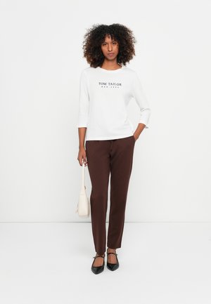 Witte shirt met "TOM TAILOR EST. 1962" in zwart gedrukt, gecombineerd met bruine broek. Accessoires zijn een kleine crème tas en zwarte flats.