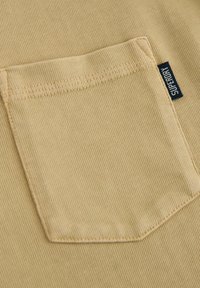 Beige bomullstyg med en strukturerad yta, som har en rektangulär bröstficka och en liten svart "Superdry"-tagg på sidan.