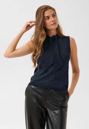 Femme aux longs cheveux ondulés portant un top sans manches bleu foncé pailleté avec un détail nœud et un pantalon en cuir noir, posant avec une main dans la poche.