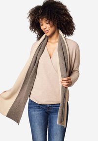 Cardigan avvolgente beige con sciarpa a contrasto grigia e crema. Texture morbida, maniche lunghe e vestibilità rilassata. Indossato sopra un top chiaro e jeans.