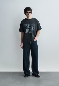 Uomo con capelli ricci che indossa occhiali da sole neri, una t-shirt nera oversize con la stampa "999 BABY", jeans ampi scuri e scarpe nere, in piedi contro un muro semplice.
