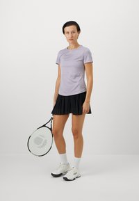 Lilla idrettskjorte med korte ermer, kombinert med et svart plisserte skjørt. Hvite joggesko og tennisracket er inkludert. Flat bakgrunn.