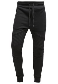 Černé joggingové kalhoty vyrobené z měkké tkaniny. Přiléhavý design s žebrovanou texturou na kolenou a dvěma bočními kapsami na zip. Nastavitelný pásek na stažení.