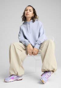 Porte-hoodie bleu clair avec cordon de serrage, pantalon large beige et baskets dégradées violettes. Coupe décontractée, texture douce et branding minimal.