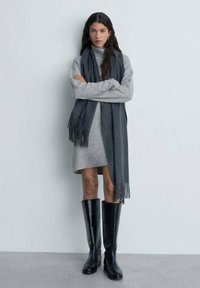 Robe en tricot grise avec une écharpe gris foncé à franges. Bottes en cuir noires jusqu'aux genoux. Fond uni.
