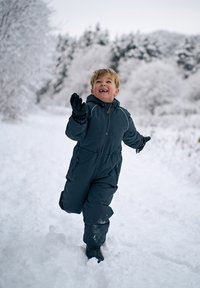 Barn i en mörkblå isolerad overall, springer genom djup snö. Har elastiska muddar och en dragkedja framtill, med snöiga omgivningar.