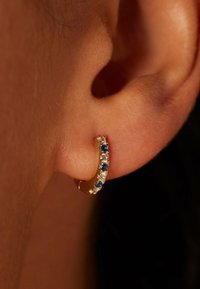 Gouden oorbel met afwisselend blauwe en heldere stenen. Gladde textuur, klein formaat, en geplaatst in het oor, waarbij het innerlijke detail zichtbaar is.