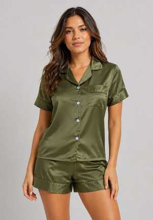 Femme debout portant une chemise en satin vert olive à manches courtes boutonnée et un short assorti sur un fond gris uni.