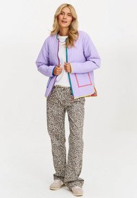 Jasnofioletowa pikowana kurtka z wielokolorową lamówką, noszona na białej koszuli, w połączeniu z beżowymi spodniami w panterkę i jasnym obuwiem.