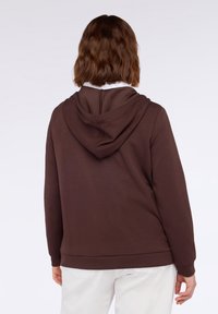 Felpa marrone con cappuccio, dalla consistenza morbida, dotata di tasca a marsupio e polsini a coste. Il retro presenta un dettaglio di cucitura e una vestibilità rilassata.