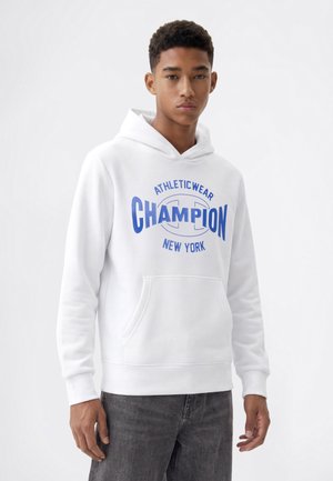 Jeune homme portant un sweat à capuche blanc avec un texte bleu "Champion Athleticwear New York" et un jean gris foncé sur un fond clair uni.