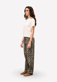 Jeune femme aux longs cheveux ondulés se tient de côté, portant un T-shirt blanc uni, un pantalon à imprimé léopard et des chaussons en fausse fourrure marron sur un fond blanc.